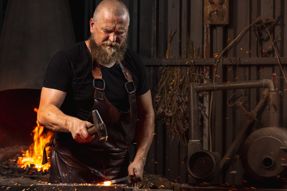 Bearded man, blacksmith manually forging the molten metal on the anvil in smithy with spark